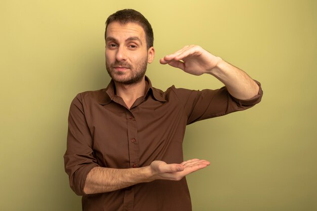 Confident young caucasian man looking at camera doing size gesture isolated on olive green background