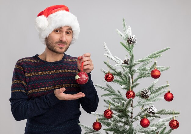 自信を持って若い白人男性がクリスマスツリーの近くに立って、白い背景で隔離のカメラを見てクリスマス安物の宝石を手で指しています。