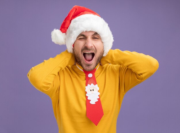 Confident young caucasian man wearing christmas hat and tie keeping hands behind neck looking at camera winking with open mouth isolated on purple background