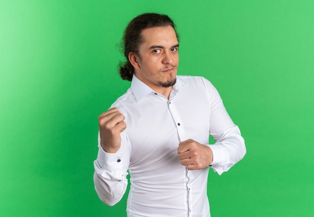 Confident young caucasian man in white shirt keeping fists ready to punch isolated on green wall with copy space