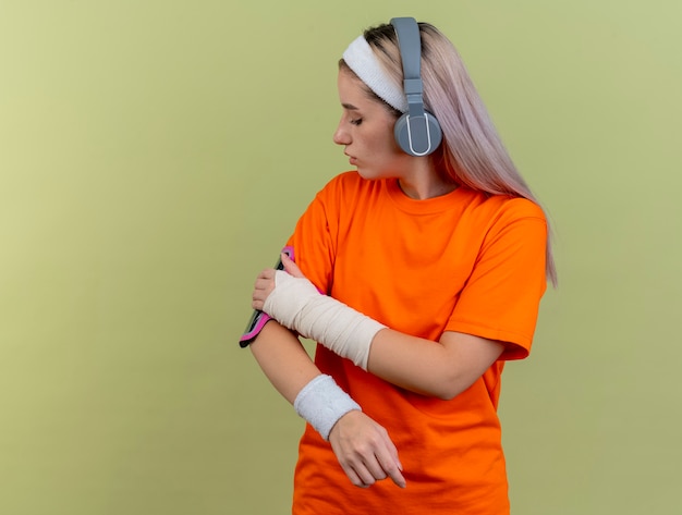 Foto gratuita fiduciosa giovane ragazza sportiva caucasica sulle cuffie che indossa fascia e braccialetti guarda e mette la mano sul bracciale del telefono