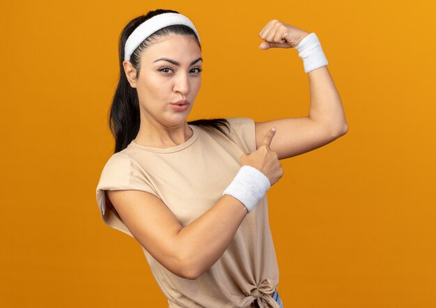 Confident young caucasian sporty girl wearing headband and wristbands standing in profile view looking at front doing strong gesture pointing at muscles isolated on orange wall