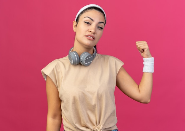 Confident young caucasian sporty girl wearing headband and wristbands with headphones around neck looking at front pointing behind isolated on pink wall