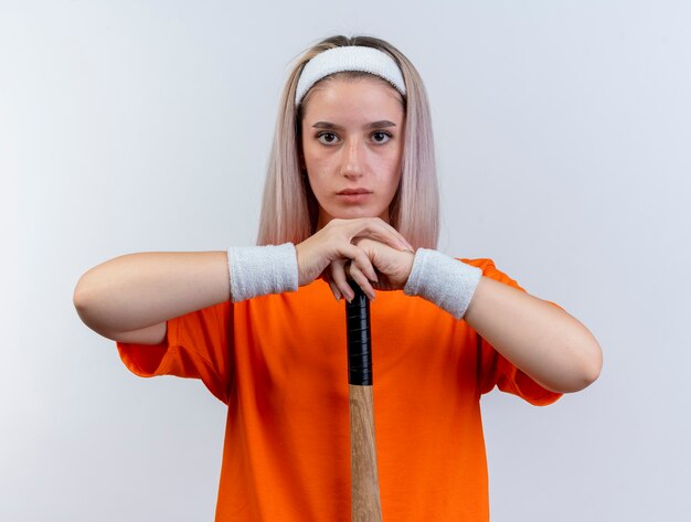 Free photo confident young caucasian sporty girl with braces wearing headband and wristbands holds baseball bat upside down