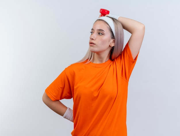 Confident young caucasian sporty girl with braces wearing headband and wristbands holds dumbbell behind head looking at side 