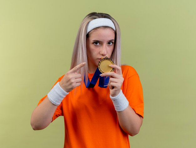 Confident young caucasian sporty girl with braces wearing headband and wristbands holds and points at gold medal 
