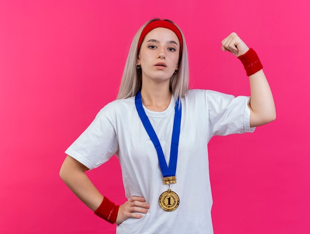 Free photo confident young caucasian sporty girl with braces and with gold medal around neck wearing headband and wristbands tenses biceps