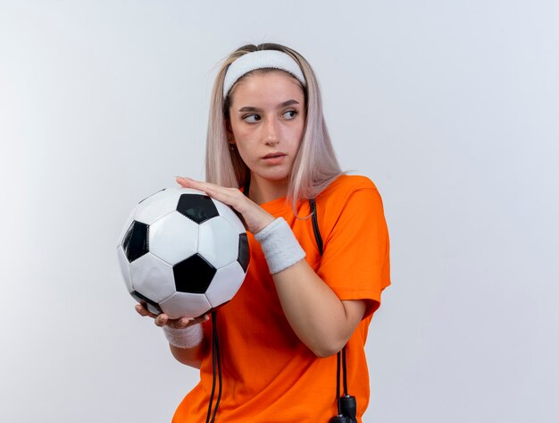 Confident young caucasian sporty girl with braces and with jumping rope around neck wearing headband and wristbands holds ball looking at side isolated on white wall with copy space