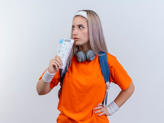 Confident young caucasian sporty girl with headphones around neck wearing backpack headband and wristbands holds air tickets looking at side 