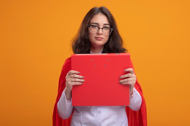 Confident young caucasian superhero girl in red cape wearing doctor uniform and stethoscope with glasses holding folder  isolated on orange wall with copy space