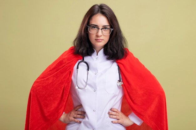 Confident young caucasian superhero girl in red cape wearing doctor uniform and stethoscope with glasses keeping hands on waist 