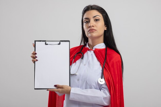 Confident young caucasian superhero girl wearing stethoscope showing clipboard looking at camera isolated on white background with copy space