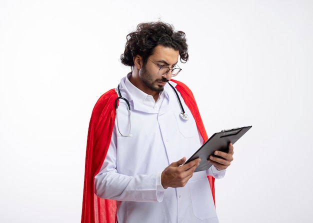 Confident young caucasian superhero man in optical glasses wearing doctor uniform with red cloak and with stethoscope around neck holds and looks at clipboard isolated on white wall