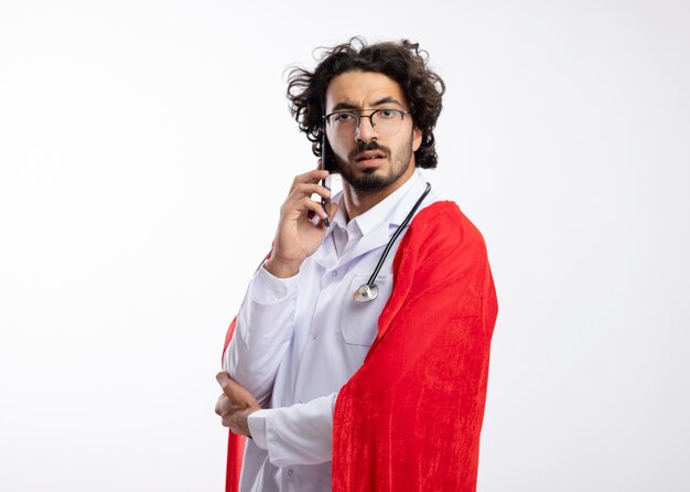 Confident young caucasian superhero man in optical glasses wearing doctor uniform with red cloak and with stethoscope around neck stands sideways talking on phone