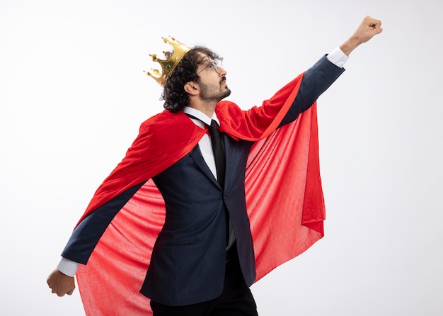 Confident young caucasian superhero man in optical glasses wearing suit with red cloak and crown stands sideways holds arms open looking and raising fist up isolated on white wall