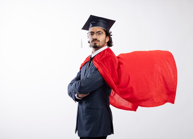 Confident young caucasian superhero man in optical glasses wearing suit with red cloak and graduation cap stands sideways 