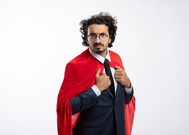 Confident young caucasian superhero man in optical glasses wearing suit with red cloak holds cloak 