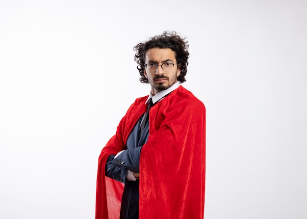 Confident young caucasian superhero man in optical glasses wearing suit with red cloak stands with crossed arms 