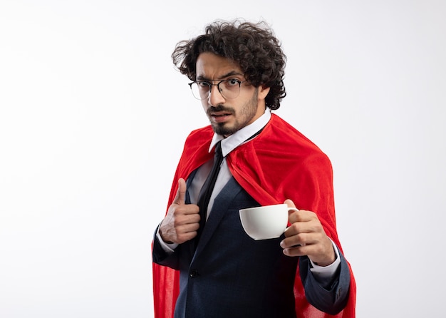 Уверенный молодой кавказский супергерой в оптических очках в костюме с красным плащом показывает палец вверх и держит чашку