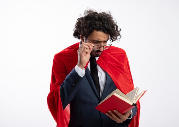 Уверенный молодой кавказский супергерой в костюме с красным плащом держит и смотрит на книгу через оптические очки