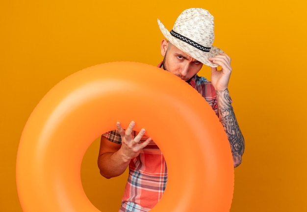 Free photo confident young caucasian traveler man with straw beach hat holding swim ring isolated on orange background with copy space