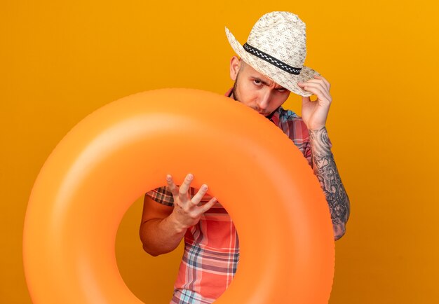 Confident young caucasian traveler man with straw beach hat holding swim ring isolated on orange background with copy space