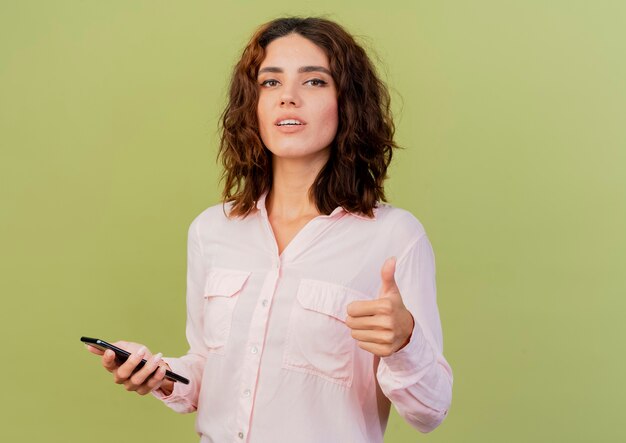 Confident young caucasian woman holds phone and thumbs up isolated on green background with copy space