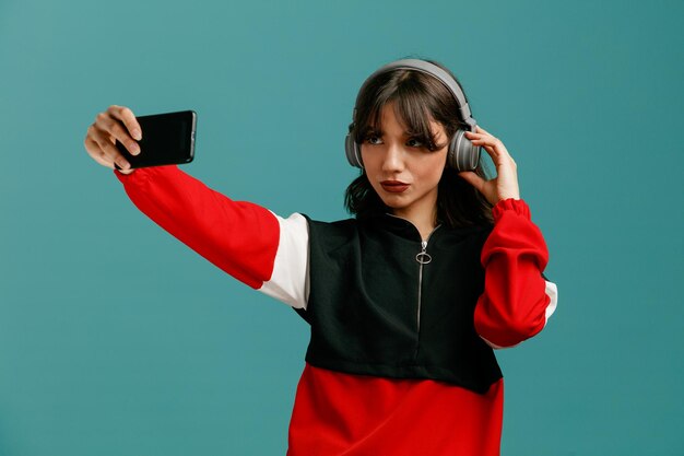 Confident young caucasian woman wearing and grabbing headphones stretching mobile phone out taking selfie isolated on blue background