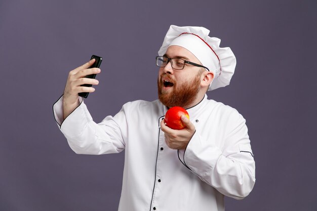 Confident young chef wearing glasses uniform and cap holding apple and mobile phone taking selfie isolated on purple background