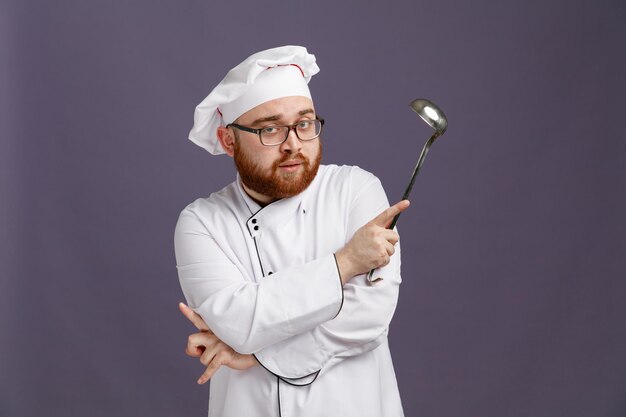 Confident young chef wearing glasses uniform and cap holding ladle looking at camera while keeping arms crossed isolated on purple background