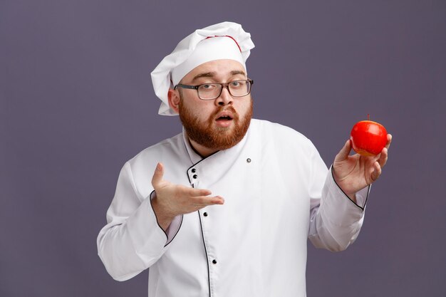 Уверенный молодой шеф-повар в очках и кепке смотрит в камеру, показывающую яблоко, указывающее на него рукой, изолированной на фиолетовом фоне