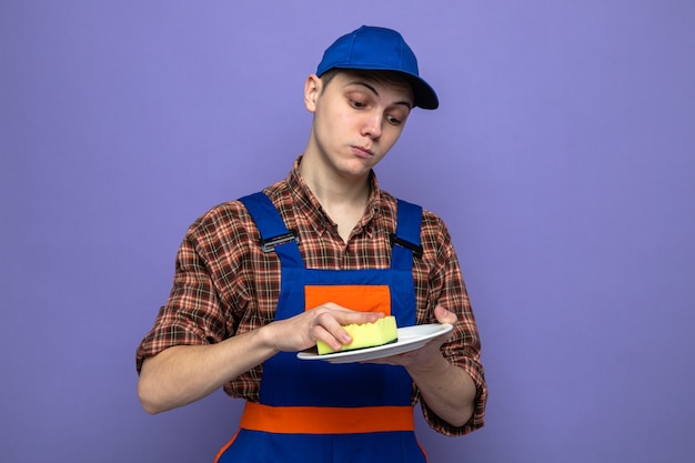 Confident young cleaning guy wearing uniform and cap washing plate with sponge 