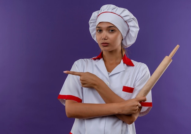 麺棒を持ってシェフの制服を着た自信のある若い料理人の女性は、コピースペースのある孤立した壁に指を向けます