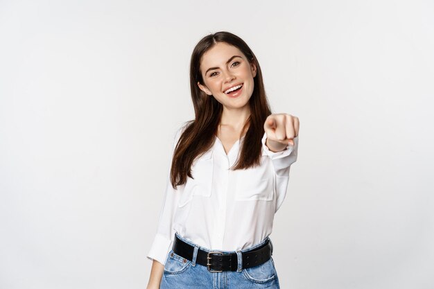 Уверенная молодая корпоративная женщина показывает пальцем на камеру, приглашает, выбирает, поздравляет, стоит на белом фоне