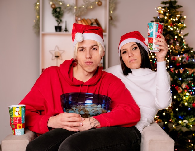 Confident young couple at home at christmas time wearing santa hat sitting on armchair guy holding bowl of potato chips girl holding plastic christmas cup both looking at camera in living room