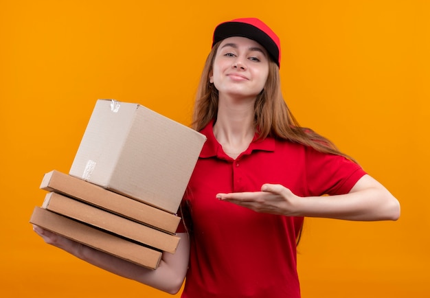 Уверенная молодая доставщица, держащая коробку и пакеты, указывающие на них рукой в красной форме на изолированном оранжевом пространстве