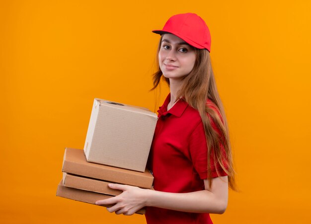 Confident young delivery girl holding box and packages standing in profile view in red uniform on isolated orange space