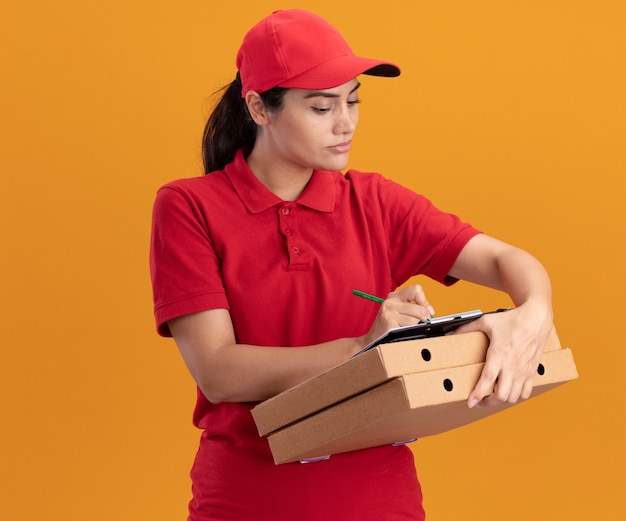 オレンジ色の壁に隔離されたピザの箱のクリップボードに何かを書く制服と帽子を身に着けている自信を持って若い配達の女の子
