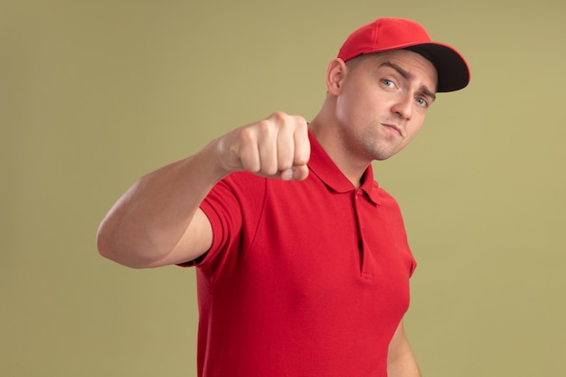 Free photo confident young delivery man wearing uniform and cap holding out fist at camera isolated on olive green wall