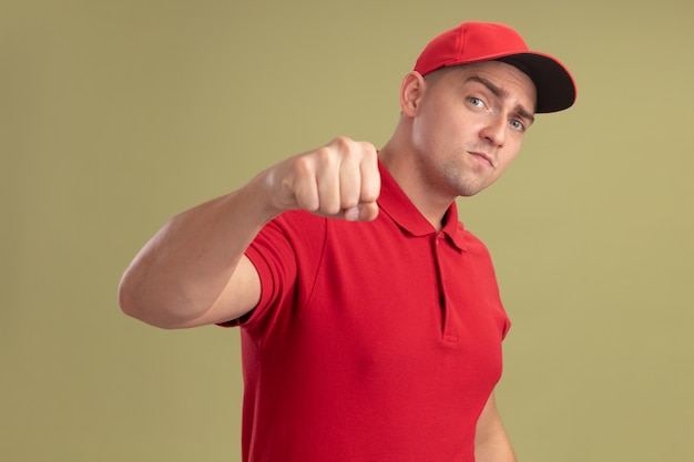 Confident young delivery man wearing uniform and cap holding out fist at camera isolated on olive green wall