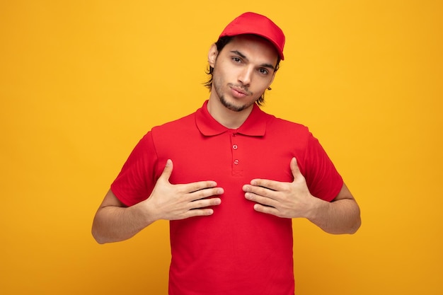 confident young delivery man wearing uniform and cap looking at camera pointing at himself with hands isolated on yellow background