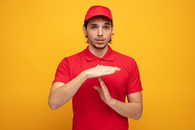 confident young delivery man wearing uniform and cap looking at camera showing timeout gesture isolated on yellow background