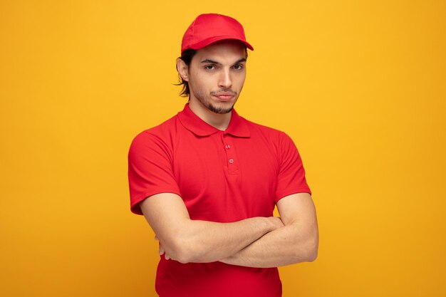 confident young delivery man wearing uniform and cap looking at camera while keeping arms crossed isolated on yellow background