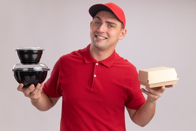 Fiducioso giovane uomo di consegna che indossa l'uniforme con cappuccio che tiene il pacchetto di cibo di carta con contenitore per alimenti isolato sulla parete bianca