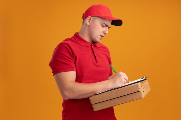 ピザの箱を保持し、オレンジ色の壁に分離されたクリップボードに何かを書くキャップと制服を着て自信を持って若い配達人