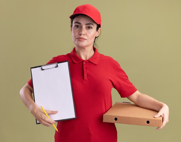 Уверенная молодая женщина-доставщик в униформе и кепке, держащая упаковку пиццы, показывает буфер обмена с карандашом в руке, изолированной на оливково-зеленой стене