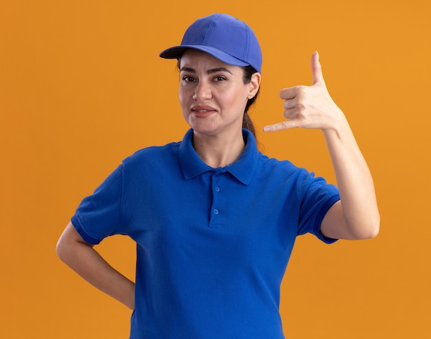 Confident young delivery woman in uniform and cap keeping hand behind back  doing hang loose gesture isolated on orange wall