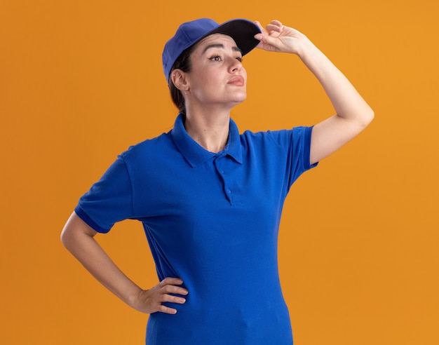 Confident young delivery woman in uniform and cap keeping hand on waist grabbing cap looking at side isolated on orange wall