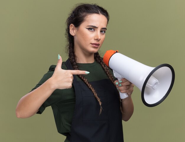 Confident young female barber in uniform holding and points at loudspeaker isolated on olive green wall