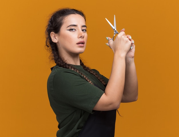 Confident young female barber in uniform holding scissors isolated on orange wall